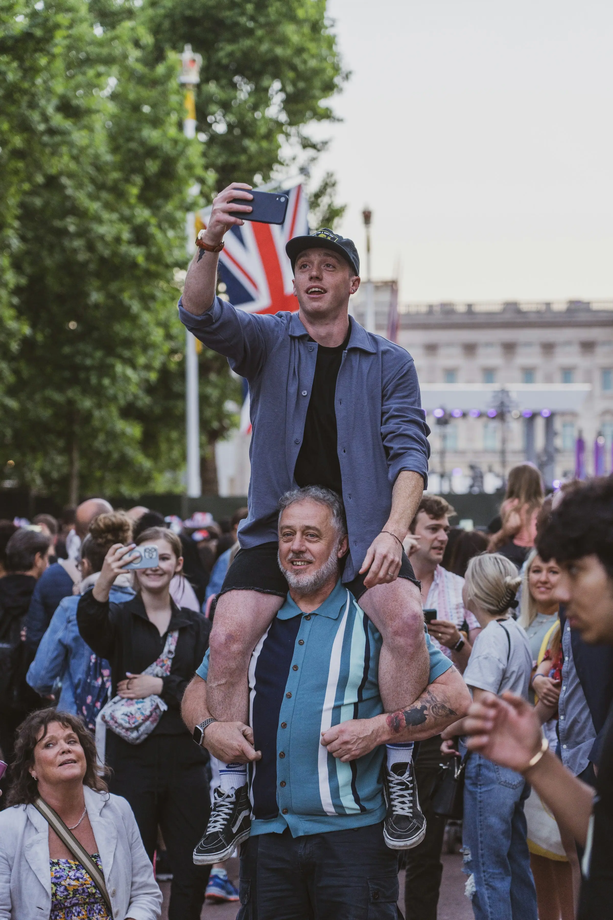 A man up on a friend's shoulders taking a photo, in a crowd on The Mall with a Union Jack behind