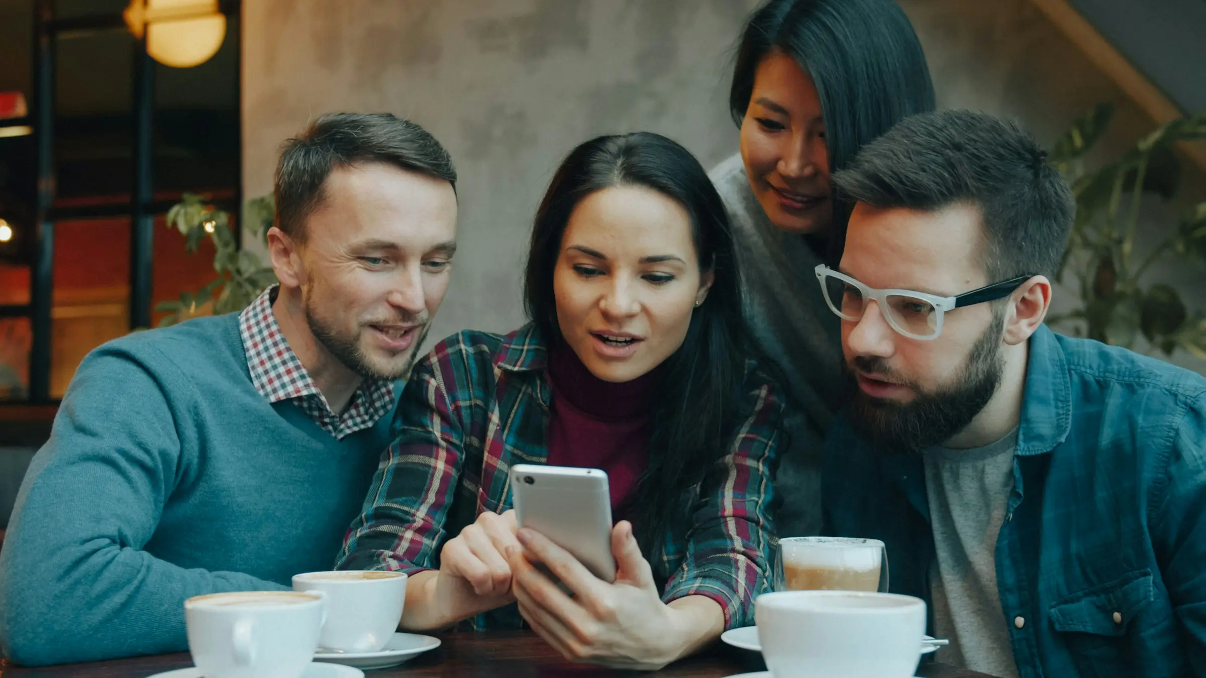 Four friends leaning in to watch something on a phone together in a café