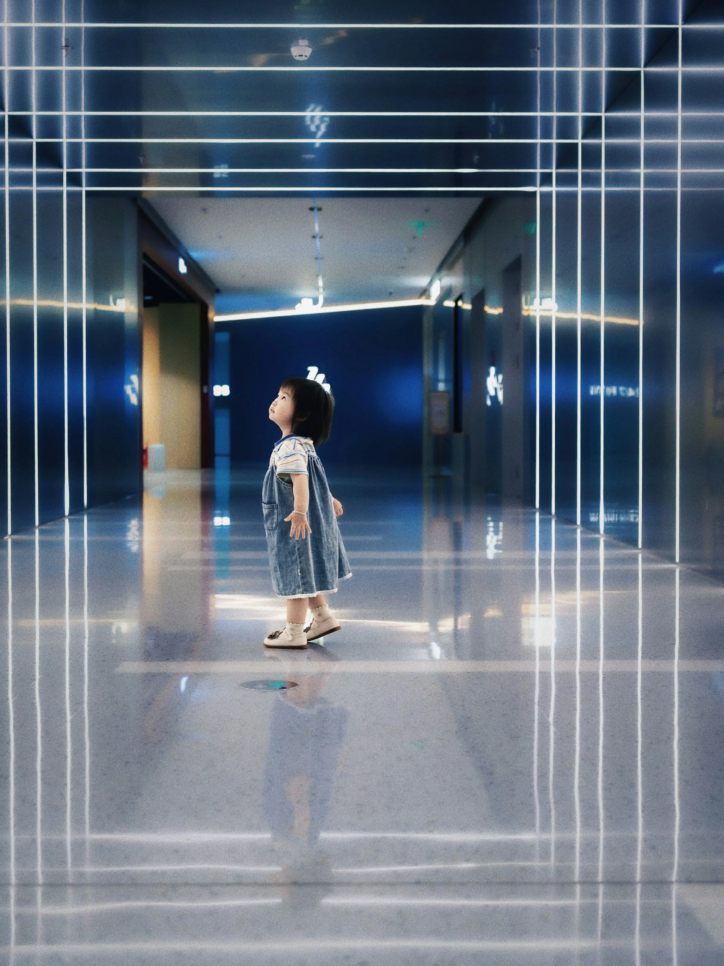 A small child standing in a long, light-filled hallway, looking up