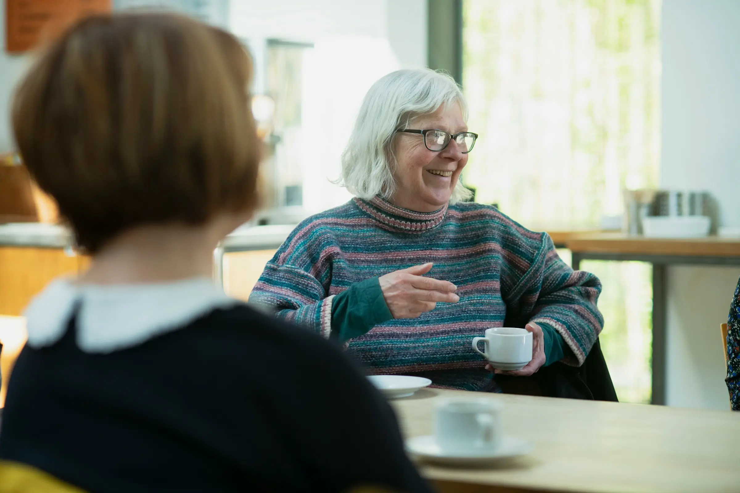 An older woman smiling as she talks over a cup of tea, listened to across the table