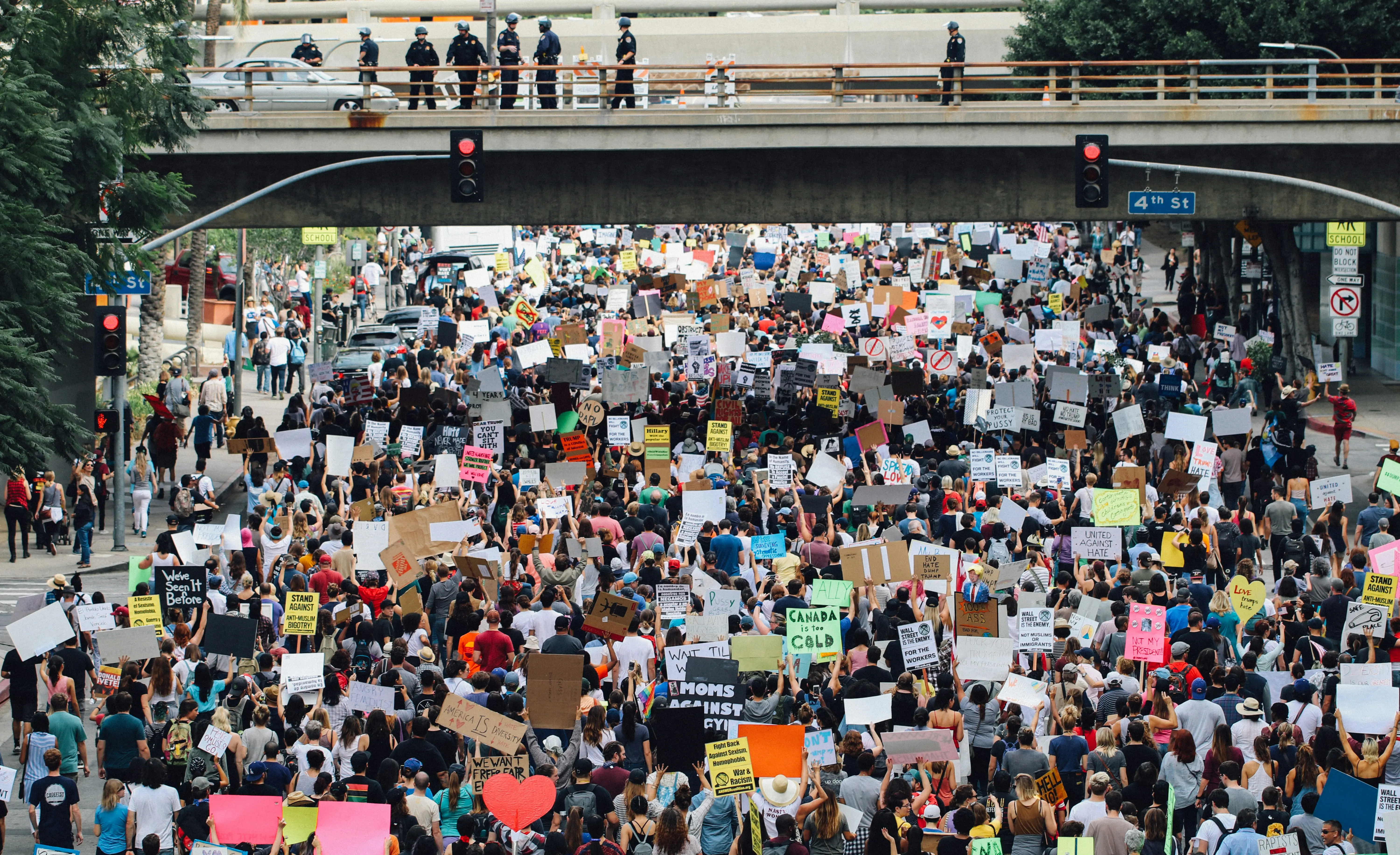 A vast crowd of marchers with placards filling a city street, police watching from a bridge above