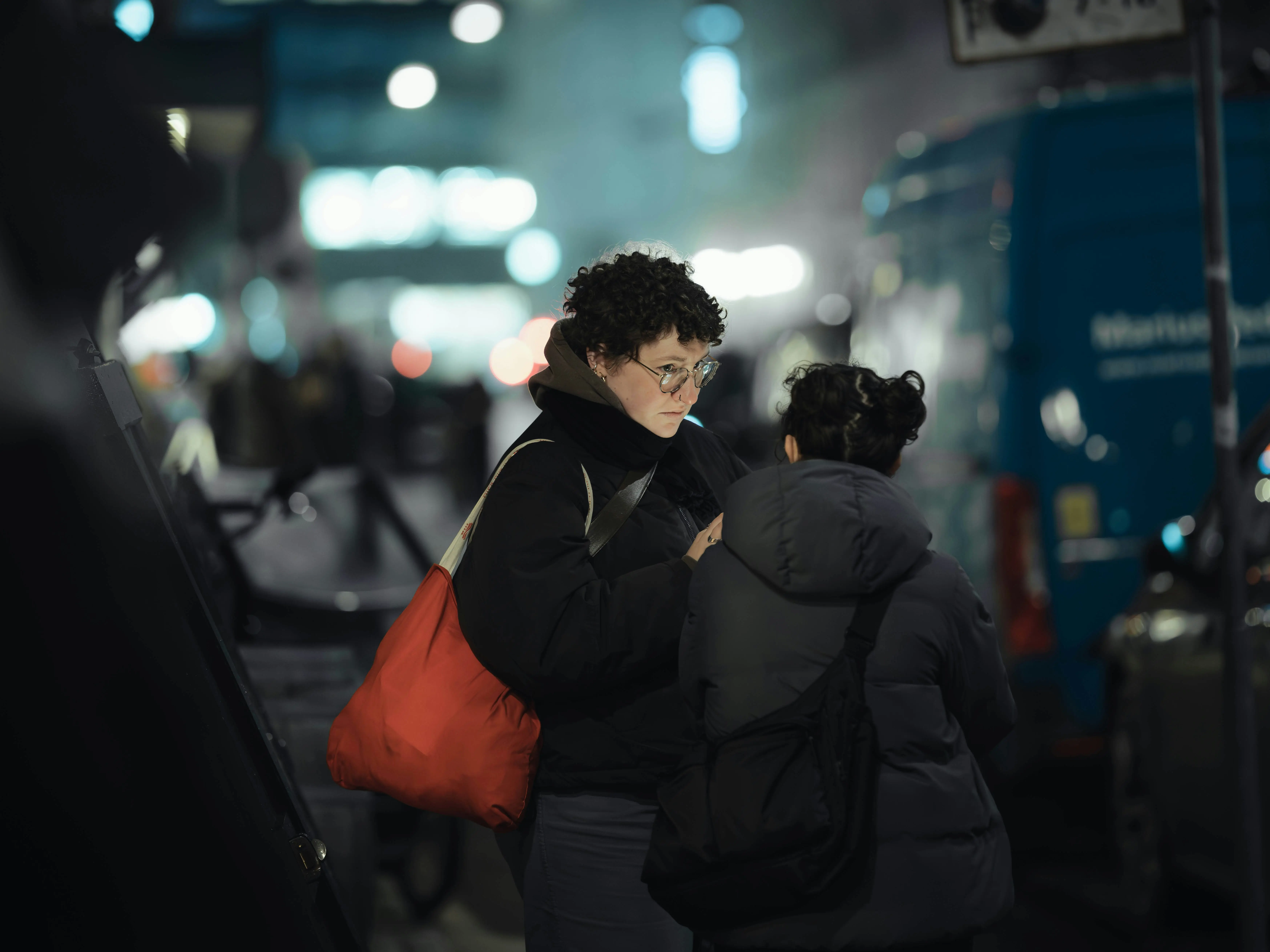 Two women talking close on a city street at night, lights blurred behind them